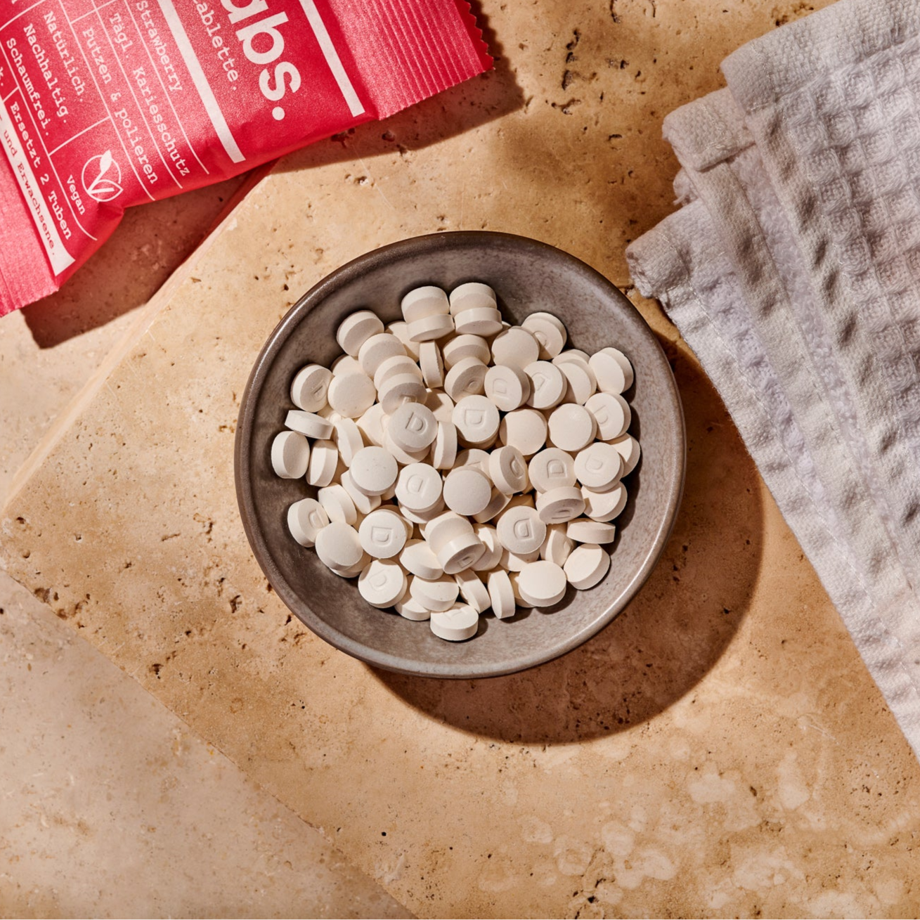 Eine kleine Schale hält weiße, runde Denttabs Zahnputztabletten auf einer hellbraunen Steinfläche neben einem gefalteten weißen Tuch und einem roten Päckchen - eine ideale Zahnpasta-Alternative.
