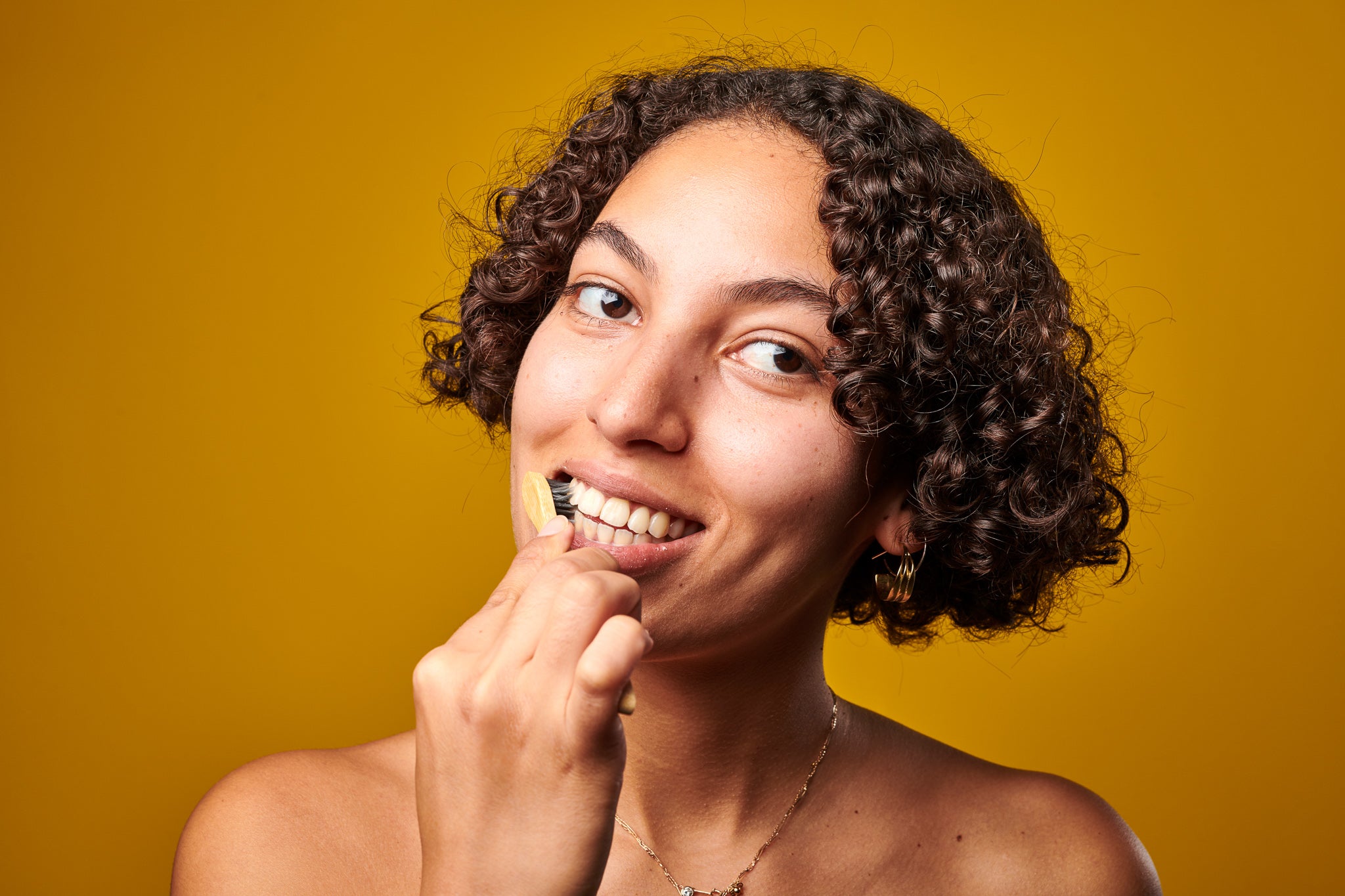 Eine junge Frau mit braunen Locken steht vor einer gelben Wand und putzt sich mit Denttabs die Zähne