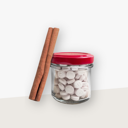 Ein Aufbewahrungsglas mit rotem Deckel gefüllt mit Denttabs in Zimtgeschmack. Eine Zimtstange lehnt an dem Glas. Der Hintergrund ist hell.